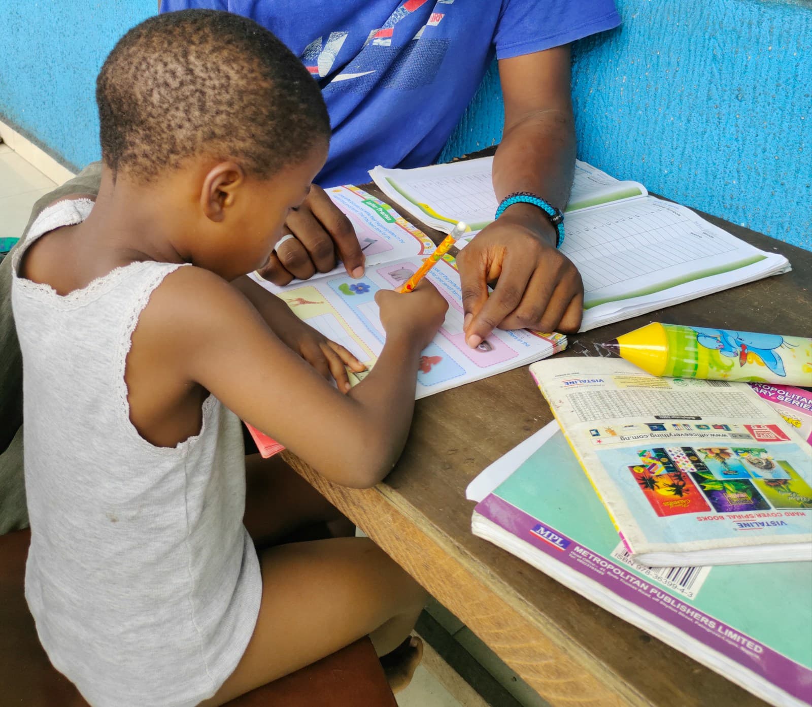 A young boy concentrates on writing in a colourful workbook while an adult beside him points to the page at a wooden table