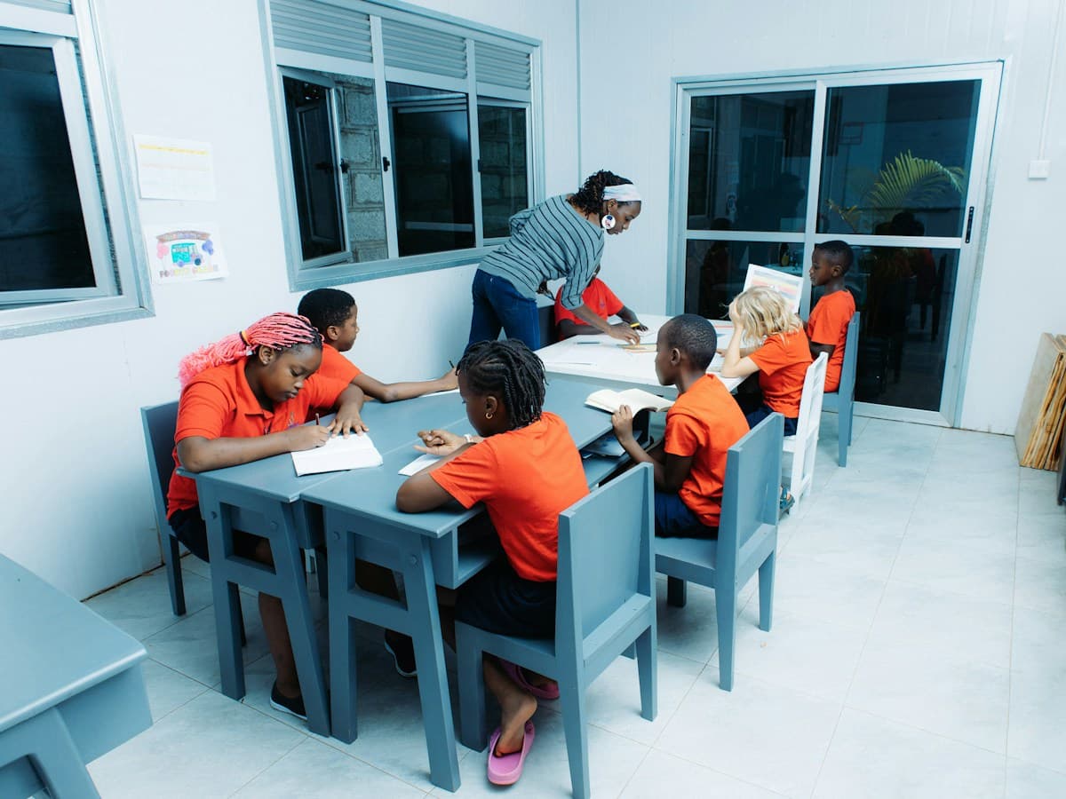 A small group of children writing at classroom tables while a teacher leans in to help one of them