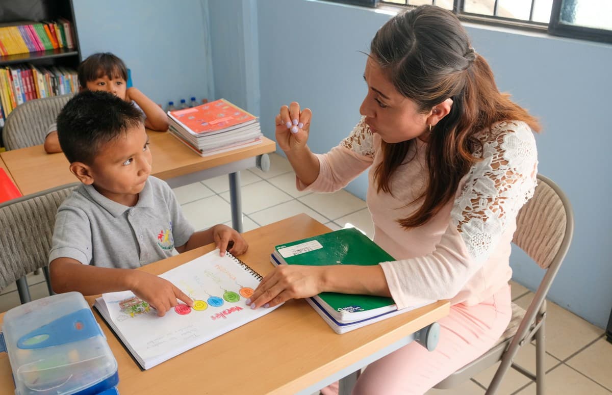A teacher sits across a desk from a young boy, pointing to a colourful page in his workbook